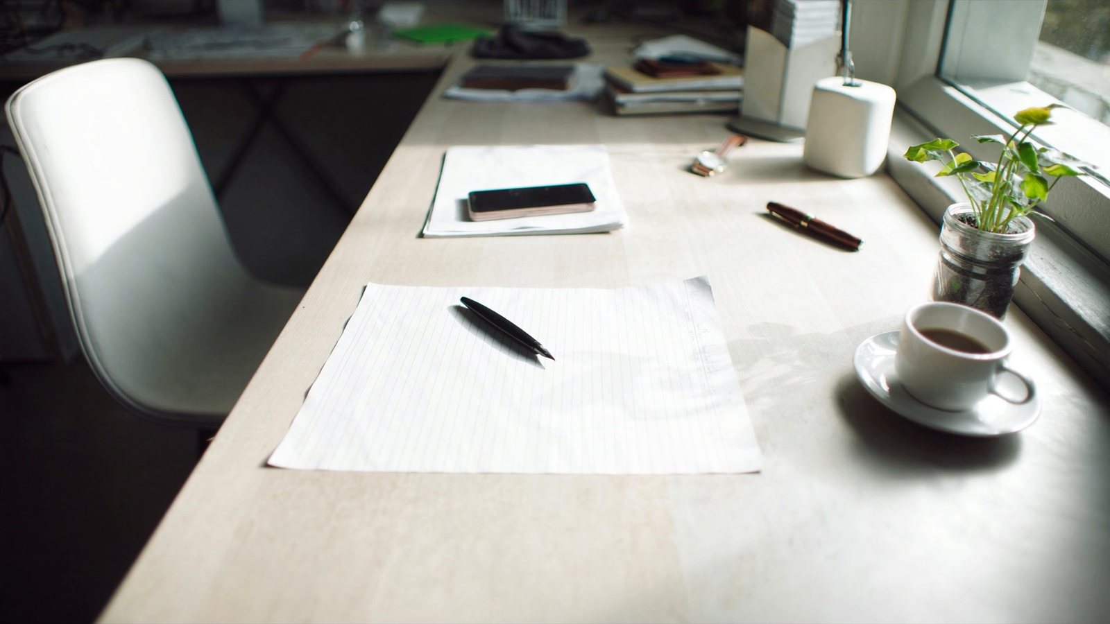 A bright desk by a window with a sheet of paper, a pen and a coffee