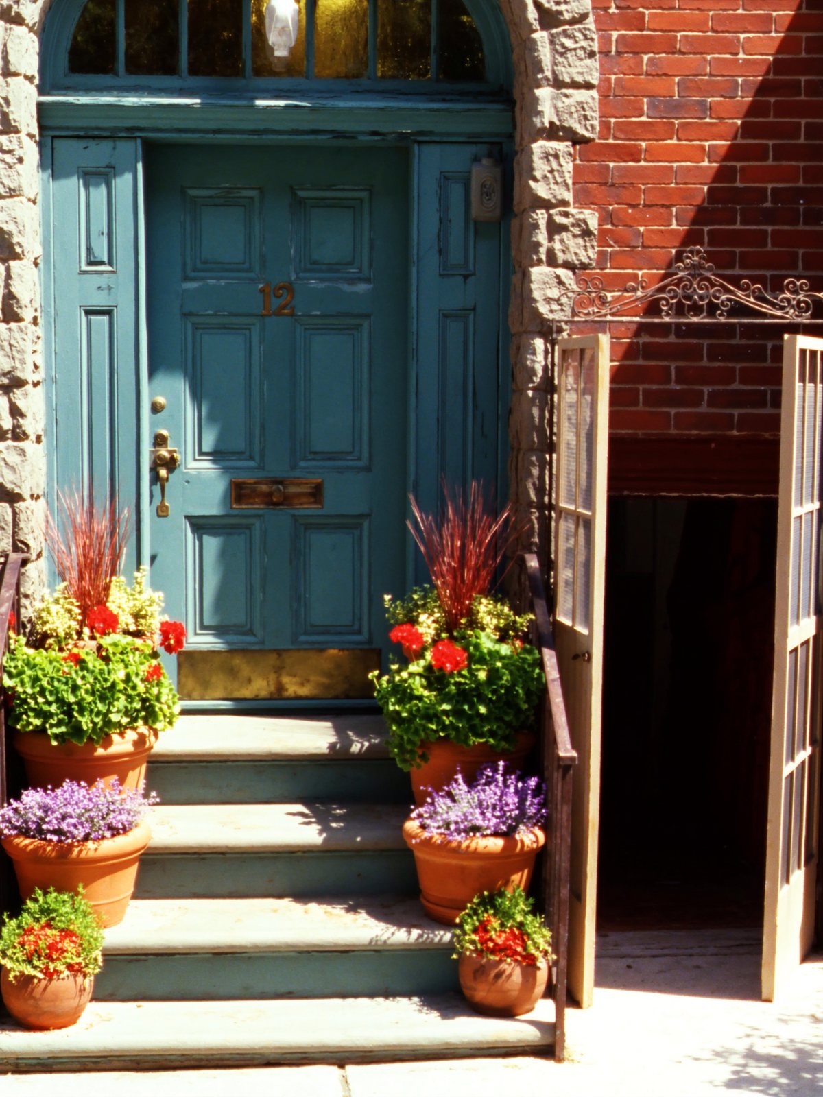 A teal front door with a brass number twelve, red geraniums on the steps