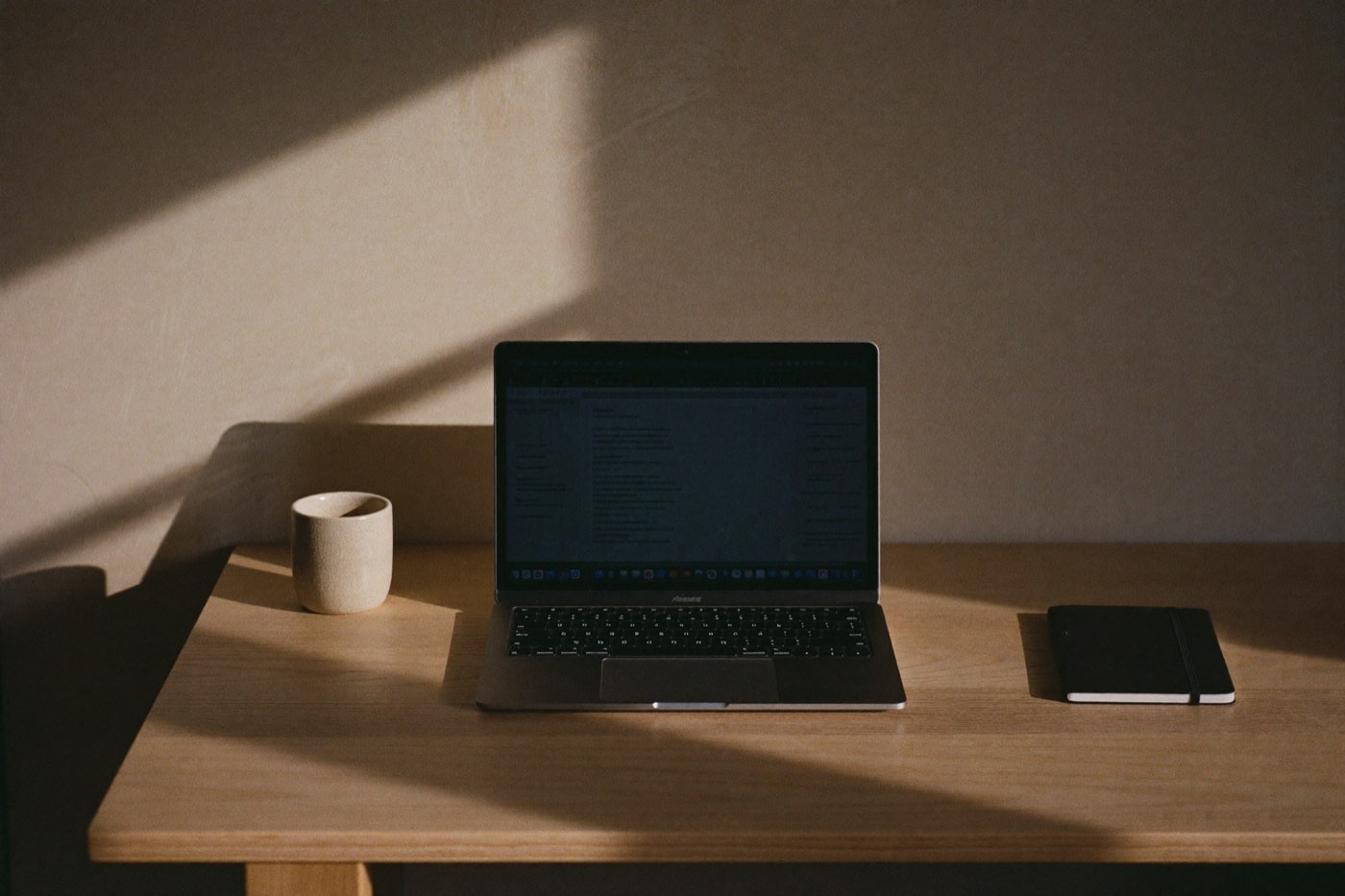 A laptop and notebook on a bare oak desk in hard afternoon light